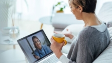 Person sitting on a couch while holding a cup and talking to a healthcare provider on their computer