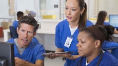 Three healthcare providers wearing scrubs sitting around a computer looking at the screen