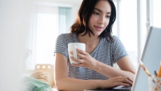 Person in a gray shirt sitting down while holding a coffee mug and looking at a computer