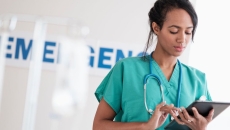 Healthcare provider wearing green scrubs and a stethoscope looking at a tablet in front of an emergency room sign