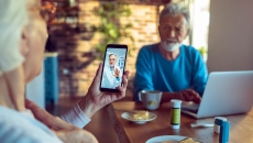 Two people sitting at a table with a computer on it and one of them is holding a phone