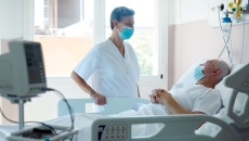 Person in mask standing over person in hospital bed