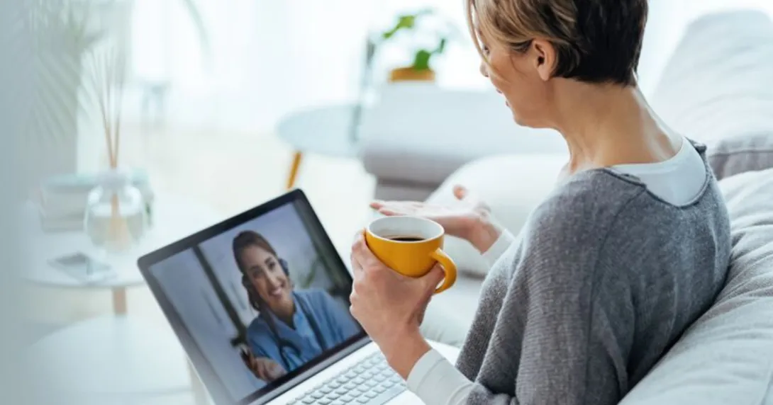 Person sitting on a couch holding a coffee cup while talking to a telehealth provider on their computer