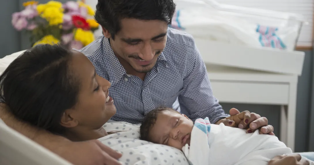 Two people, one lying in a hospital bed and the other leaning over the bed, with both looking at a baby