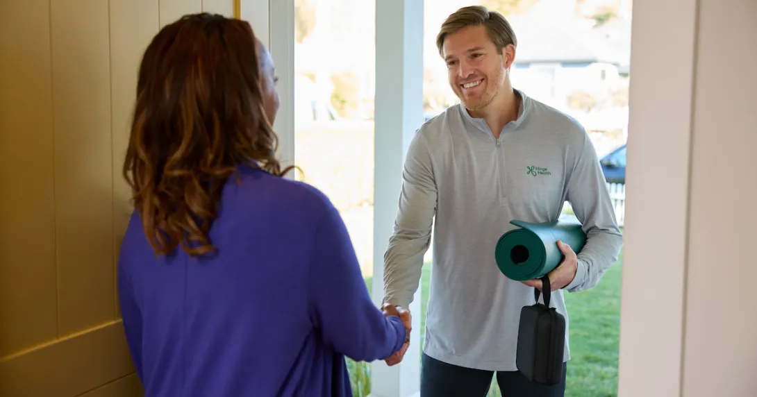A physical therapist greeting a patient at the door with a yoga mat
