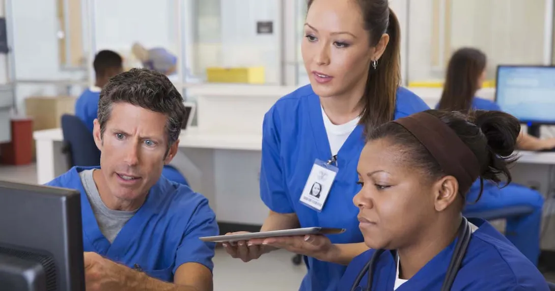 Three healthcare providers wearing scrubs sitting around a computer looking at the screen