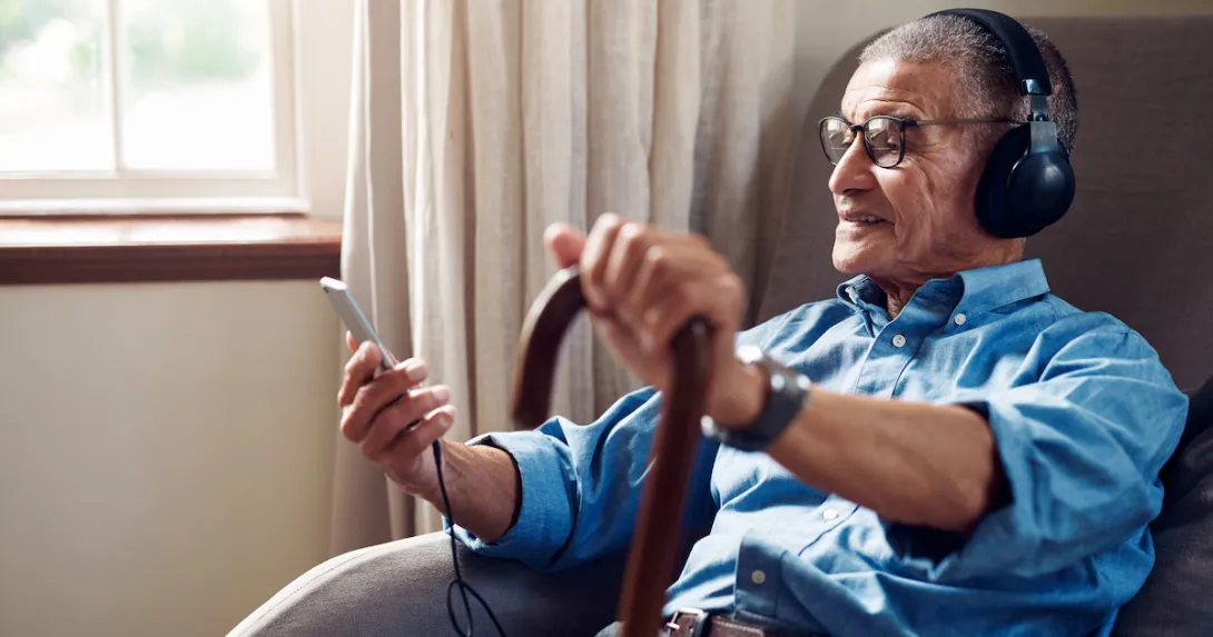 An older man in his home wearing headphones and using a smartphone.