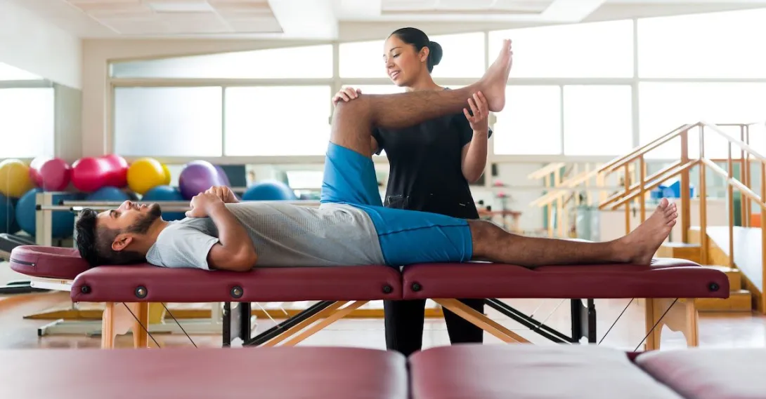 A male patient lying on his back on a massage table and a female physical therapist holding his leg up.