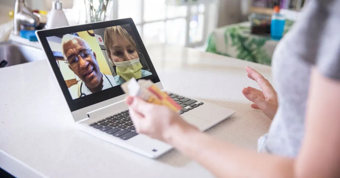 A close up of a woman holding a pill bottle while talking to two providers on her laptop.