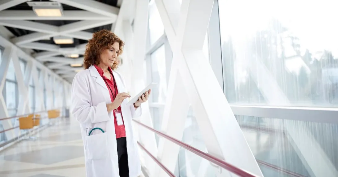 Healthcare provider holding a tablet while wearing a lab coat and standing in a hallway by a large window