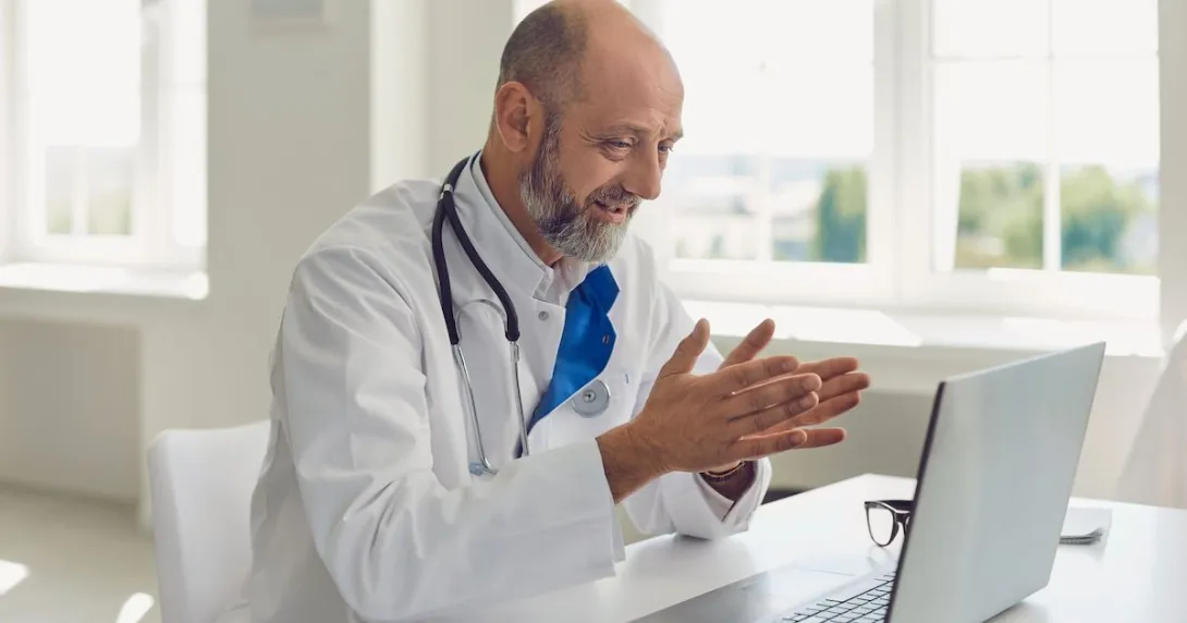 Healthcare provider sitting at a desk, wearing a lab coat and stethoscope, while looking at a computer