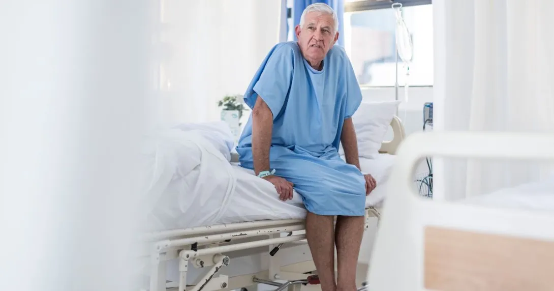 Patient sitting on the side of a bed in a hospital room wearing a hospital gown