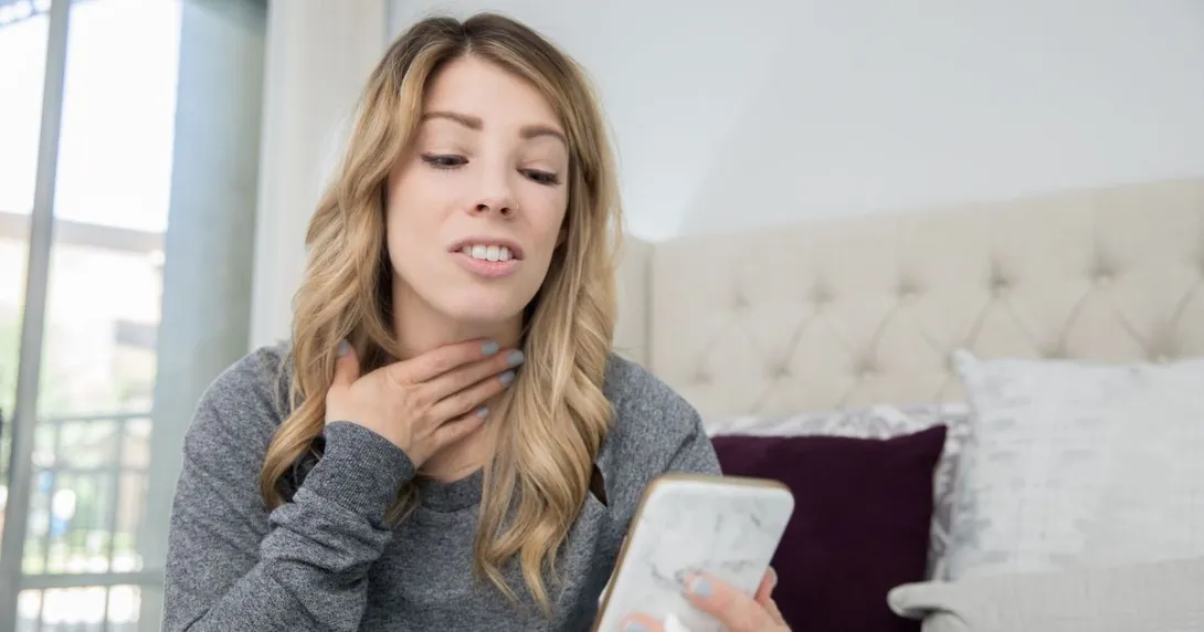 A woman rubbing her sore throat holding a smartphone.